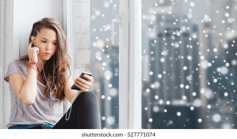 People, Technology, Winter, Christmas And Teens Concept - Sad Pretty Teenage Girl Sitting At Window With Smartphone And Earphones Listening To Music Over City Background Over Snow