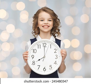 People, Time Management And Children Concept - Smiling Girl Holding Big Clock Showing 8 O'clock Over Holidays Lights Background