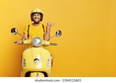 People, transport and advertisement concept. Cheerful young European male motorcyclist points thumb away, dressed casually, poses on own motorbike, glad to show copy space for your information