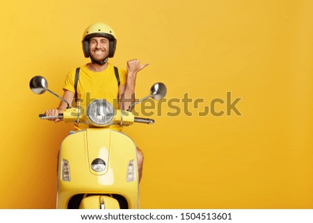 People, transport and advertisement concept. Cheerful young European male motorcyclist points thumb away, dressed casually, poses on own motorbike, glad to show copy space for your information