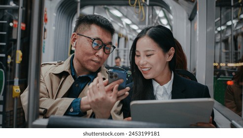 People traveling together at night in train or bus. Sitting down while using digital devices. Man showing funny video to woman. Asian girl smiling and replying back. Man can't hear through earphones. - Powered by Shutterstock