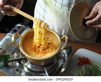 People using chopsticks to cook ramyeon or Korean instant noodles soup in double handle Korean yellow aluminum pot on gas stove over wooden table with slided red chili, scallion on cutting board.