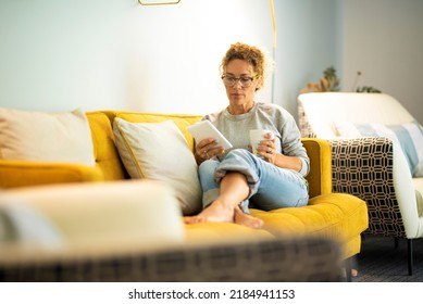 People Using Electronic Device For Relax And Leisure Activity Indoor At Home. One Woman Comfortably Sitting On A Yellow Couch Reading E-book From Modern Reader. Female Enjoying Time To Study Online