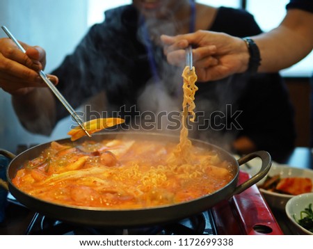 People using silver chopsticks to pick up noodles and vegetable from hot pot of budae jjigae or Korean stew made with kimchi, instant noodle (ramyeon), ham, sausage, tofu, vegetables / selective focus