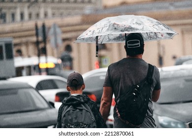 People Are Waiting For A Taxi In The Rain Outdoors. Family Under An Umbrella In The Downpour Waiting For The Car. Father With Son On The City Streets In Bad Weather