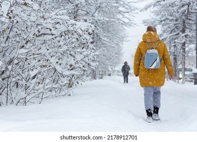 People walk on a snow-covered sidewalk during a heavy snowfall. Lots of snow on the ground and branches of trees and bushes. Cold snowy winter weather. Woman in warm winter clothes with a backpack.
