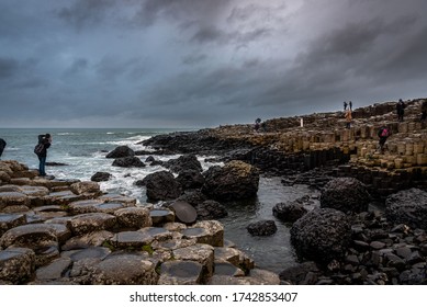 People Walking Over The Natural Hexagonal Stones At The Coast Called Giant's Causeway, A Landmark In Northern Ireland.