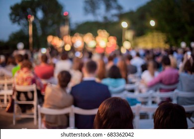 People are watching a concert of classical music. blurred young and adult women and men and other people watching listening to concert of classical band in daylight outdoor enviroment.
