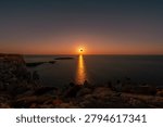 People watching a total solar eclipse at sunset from the rocky coast of Menorca with sunburst reflection on ocean