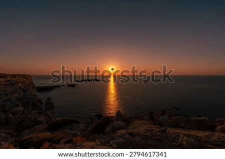 People watching a total solar eclipse at sunset from the rocky coast of Menorca with sunburst reflection on ocean