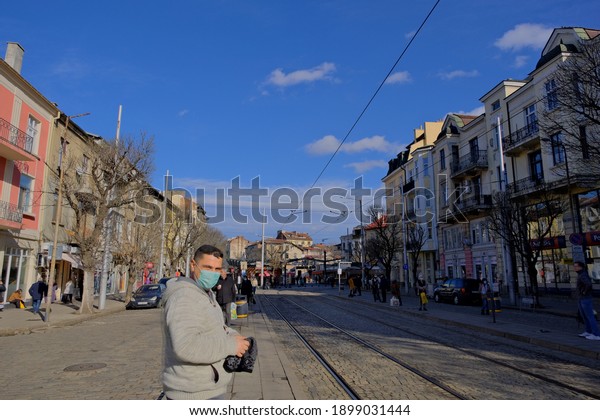 People wearing medical mask in Sofia street\
near the bazaar called as women bazaar on the cobblestone and\
people walking around and cars parking on the street. Bulgaria.\
Sofia. 06.01.2021.