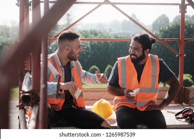 People Working In Construction Site. Men At Work In New Housing Project. Team Of Happy Workers Laughing, Talking And Eating Snack During Lunch Break