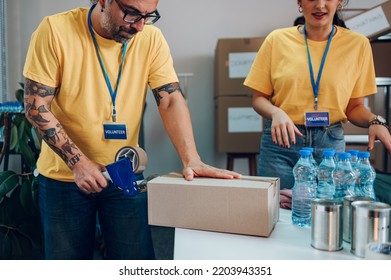 People Working On Humanitarian Aid Project. Teamwork In Homeless Shelter. Volunteers Working In A Community Charity Donation Center. Focus On A Middle Aged Man Putting Duck Tape On A Cardboard Box.