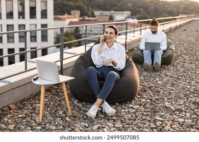 People working remotely on rooftop using laptops and bean bags. Man focused on laptop, woman talking on phone. Relaxed outdoor work environment with cityscape background - Powered by Shutterstock