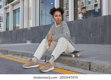 People youth generation and hobby concept. Thoughtful millennial girl with two hair buns rests at longboard dressed in streetwear enjoys recreation chill poses at urban setting. Stylish skateboarder