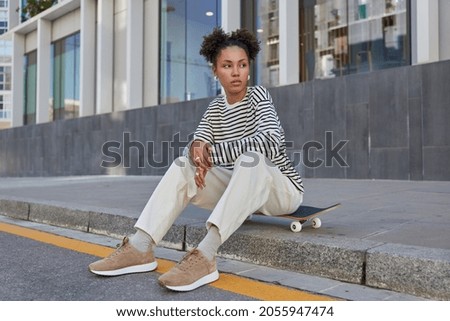 People youth generation and hobby concept. Thoughtful millennial girl with two hair buns rests at longboard dressed in streetwear enjoys recreation chill poses at urban setting. Stylish skateboarder