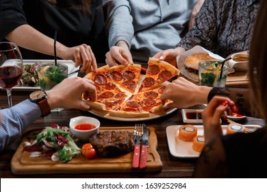 People's hands taking slices of original classic pepperoni pizza on wooden table background with other food: steak, burger, sushi rolls, salad, cocktails and wine in restaurant. Side view.