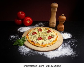 pepperoni and olive pizza, on a wooden board, is accompanied by fresh ingredients, flour, tomatoes and basil, and a salt and pepper shaker.