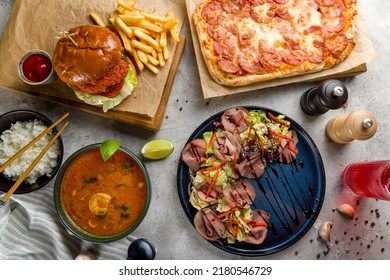 Pepperoni pizza on Roman dough pinsa, salad with roastbeef, burger with meat patty and soup tom yam with rice on grey table top view