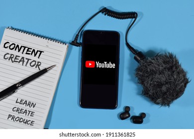Perak, Malaysia - February 6,2021 : Content Creator Wording on A Notebook, YouTube Logo on A Smartphone, Black Ear Buds And A Mic With Blue Background. People working as A Content Creators in YouTube 
