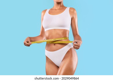 Perfect Body Concept. Cropped close up view of slim woman in white top and panties measuring her waist with yellow tape ruler. Unrecognizable lady checking waistline size isolated on blue studio wall