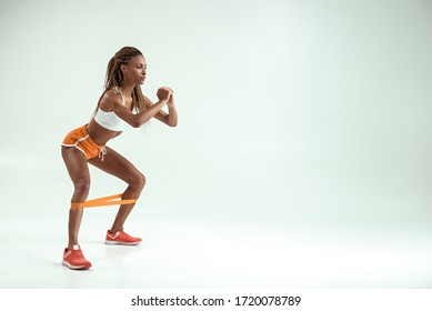Perfect buttocks. Side view of young and slim african woman in sports clothing doing squat exercises with a resistance band while standing in studio against grey background
