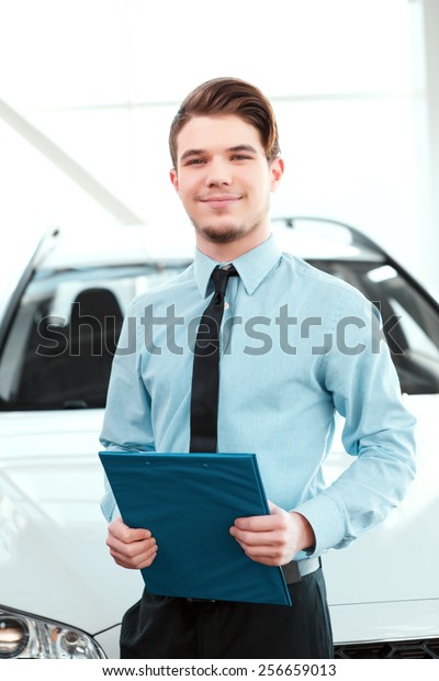 Perfect car dealer. Portrait of handsome young car sales\
man in formalwear holding a clipboard and smiling at camera in a\
car dealership 