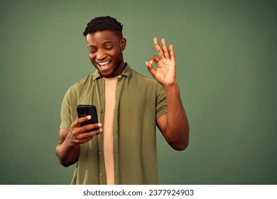 Perfect choice. African american young guy showing okay sign gesture while looking at smartphone screen with sincere smile on face. Satisfied man enjoying new mobile app over green studio background. - Powered by Shutterstock