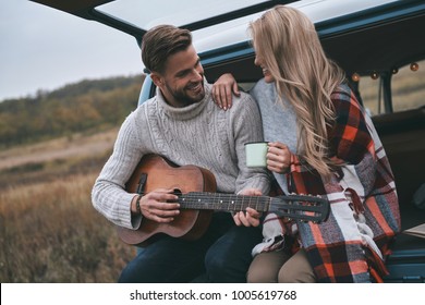 Perfect date.  Handsome young man playing guitar for his beautiful girlfriend while sitting in the trunk of blue retro style mini van - Powered by Shutterstock