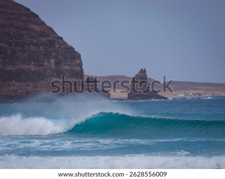 Perfect empty wave rolling beneath rugged volcanic cliffside with sea spray rising into the air. Picturesque surf spot surrounded by a rocky seascape and a lonely rock formation at the end of a cape.