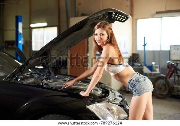 Perfect female of sports\
constitution in short shorts and a top near the open hood of a\
black car in the garage. A girl with a beautiful smile looks at the\
camera