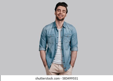Perfect man. Handsome young man in blue jeans shirt keeping hands in pockets and looking at camera while standing against grey background