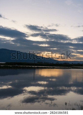 Perfect mirror reflection of dramatic evening clouds and distant mountains in a vast flooded agricultural field at dusk. The scene represents pristine natural beauty, environmental conservation, and profound peacefulness.