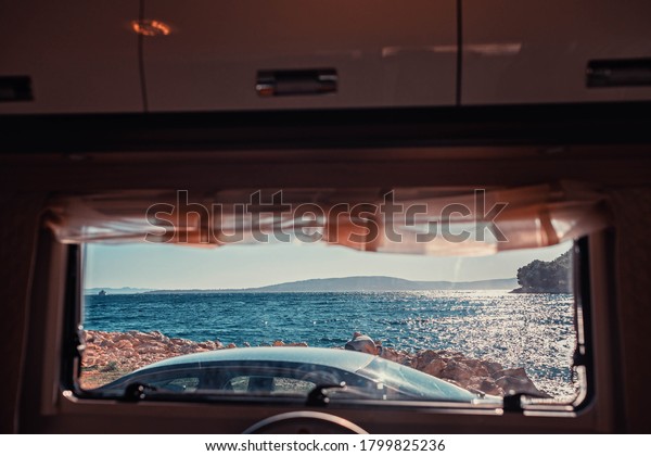 Perfect place to camp. Window is\
perfectly framing blue shining sea. Car is parked next to campervan\
on the coppery sand. There is solitary mountain at\
horizon.