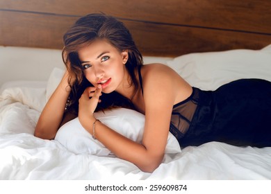 Perfect sexy tan woman with  perfect skin , big full lips posing at bedroom, wearing sexy luxury stylish  black lingerie.l Lady lying on her stomach on white bed. 