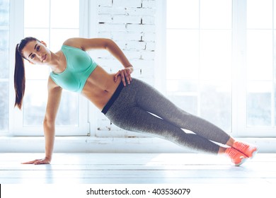Perfect side plank. Full length of young beautiful woman in sportswear doing side plank and looking at camera in front of window at gym 