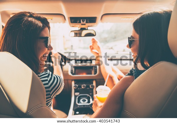 Perfect start of holidays. Rear view of two beautiful\
young cheerful women looking at each other with smile while sitting\
in car 