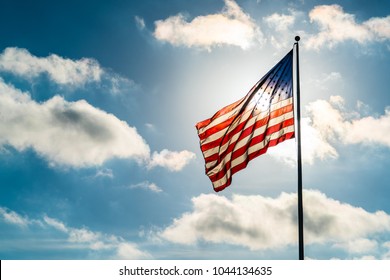 Perfect Sunny Day With Puffy White Clouds And Direct Sunshine Into The Perfect American Flag Illuminated Back Lit By Sun. United States Flag Waving In The Wind On A Dramatic Sky Day Direct Sunshine