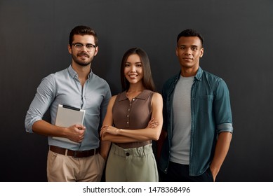 Perfect IT Team. Group Of Three Cheerful Young People In Casual Wear Looking At Camera With Smile While Standing Against Dark Background