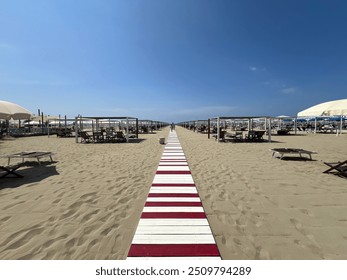 A perfectly aligned beach setup at Forte dei Marmi with a red and white walkway leading to rows of umbrellas and sunbeds. The clear blue sky and pristine sand create a luxurious and organized atmosphe - Powered by Shutterstock