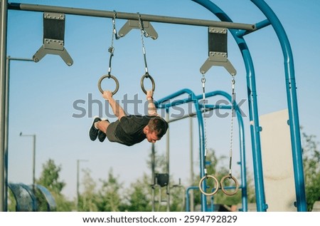 Perfectly parallel to the ground, a man confidently executes an iron cross on rings in a bright outdoor setting.
