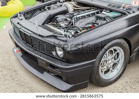 Performance car front end featuring exposed engine components, supercharger intake, clean engine bay, matte black bumper with aerodynamic design, integrated round headlight, polished five-spoke allo.