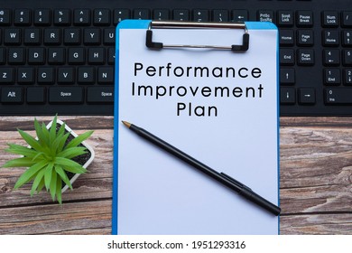 Performance Improvement Plan Text On Blue Clipboard On Top Of A Keyboard With Pen And Potted Plant On Wooden Desk