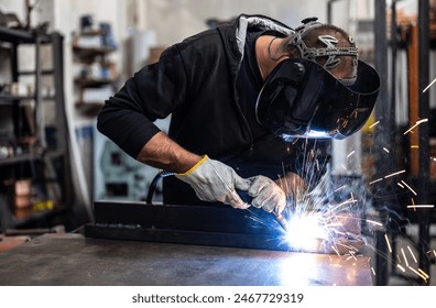 Performing a weld: the man in the welding mask is positioned with the welding machine on the welding point. When he activates it, a blue glow with smoke and orange sparks illuminates the scene. - Powered by Shutterstock