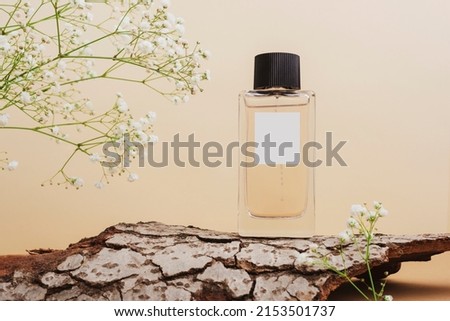 Perfume bottle with blank label on tree bark and white gypsophila flowers on neutral beige background. Front view.