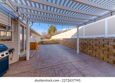 Pergola roof with stringlights above the stone tiles ground at the backdoor of a house. There is a griller near the house with glass doors and a wall with concrete blocks and fence panel on the right.