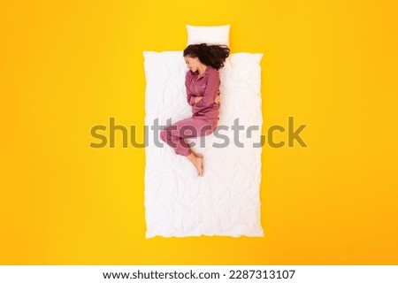 Period Pain. Unhappy Woman Suffering From Menstrual Cramps And Stomachache Sleeping Hugging Herself Over Yellow Studio Background, Wearing Pajamas. Female Health Problems. Top View