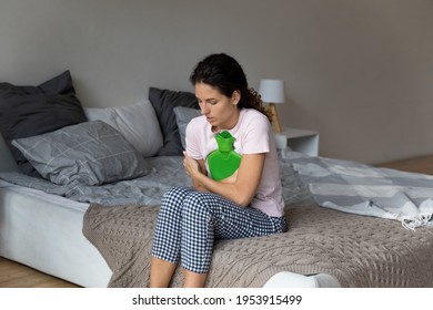 Periodical pain. Stressed hispanic lady sit on large bed with closed eyes feel stomach ache menstrual cramps diarrhea cystitis symptoms. Unhealthy young female hug warm water bag to relieve belly pain