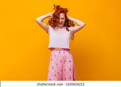 Perky Girl In Pink T-shirt And Skirt Ruffles Her Red Hair Against Orange Background