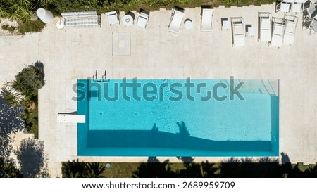 Perpendicular aerial view of a rectangular swimming pool located in the garden of a villa. The pool and sun loungers are empty. A garden surrounds the pool.
